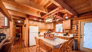 L’intérieur d’une cabane en rondins comprend une cuisine avec des meubles en bois, un réfrigérateur, un coin repas avec des tabourets de bar et une cheminée.