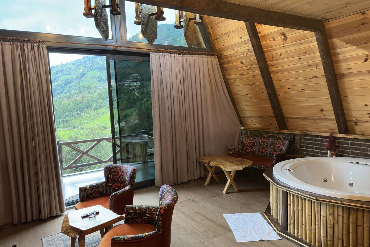 Een knus interieur met een houten plafond, een groot raam met gordijnen, een houten tafel, twee gestreept stoelen, een kleine bank en een whirlpool.