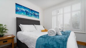 Una cama bien hecha con sábanas blancas, una manta azul y toallas enrolladas. Hay una mesita de noche de madera con una planta y una bandeja de fruta. Dos cuadros abstractos cuelgan sobre la cama.
