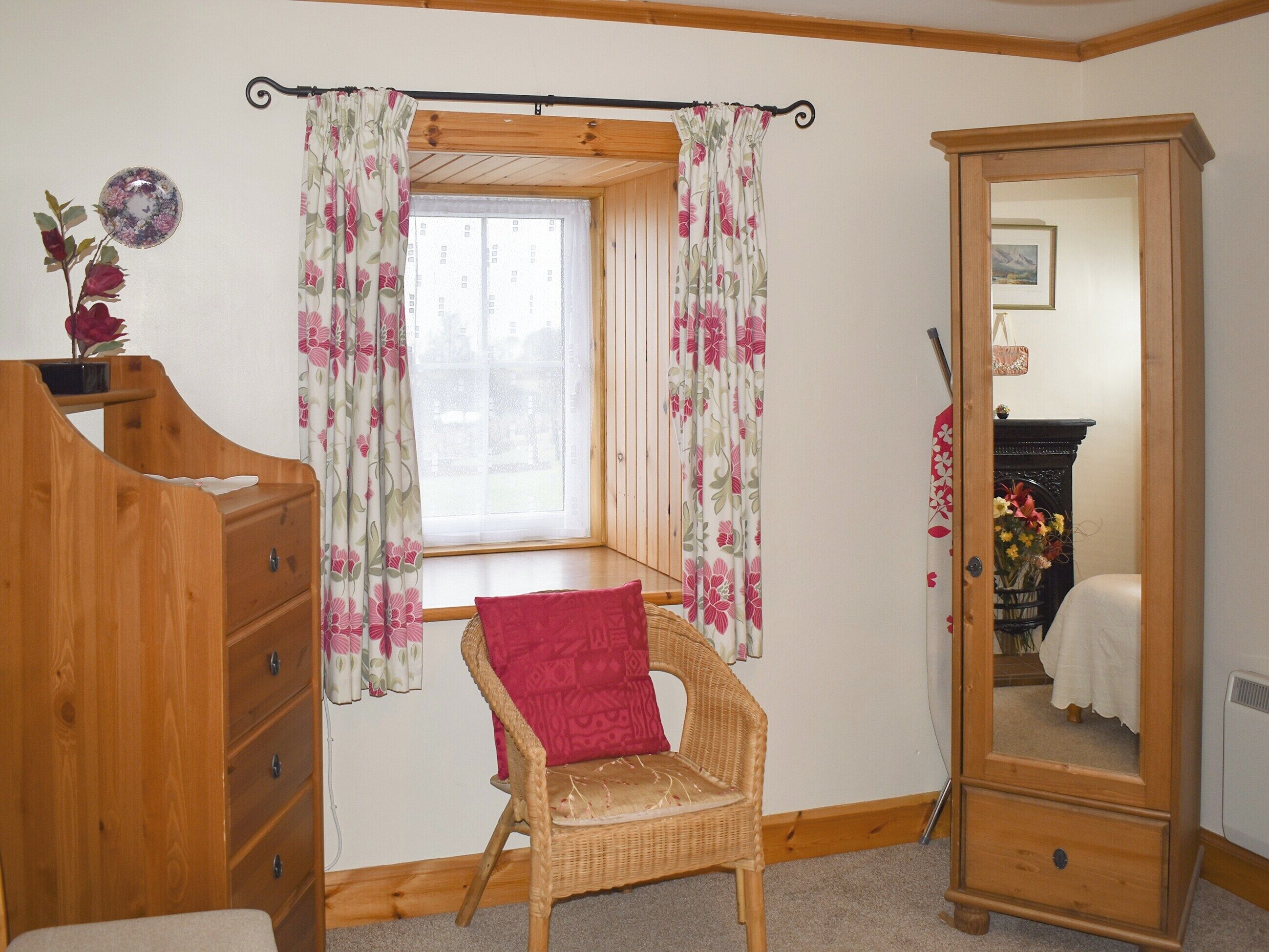 Une pièce comprenant une commode en bois, une chaise en osier avec un coussin rouge, une fenêtre ornée de rideaux à motifs floraux et un meuble avec une porte-miroir.