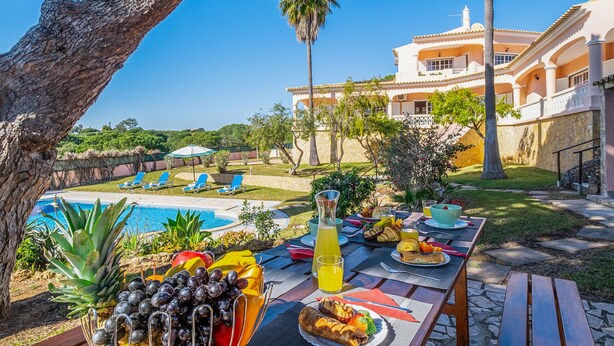 Zona de comedor junto a la piscina con una mesa dispuesta para el desayuno, con vistas a una alberca y una casa grande con jardín.