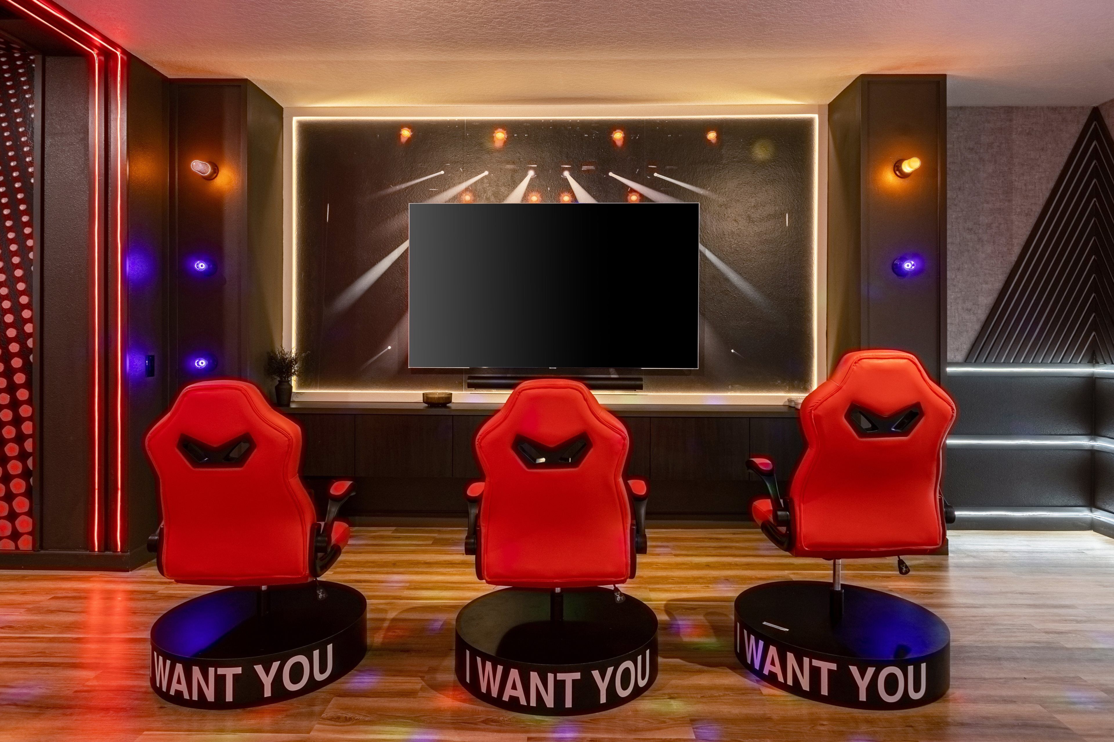 Una camera moderna con una grande TV a schermo piatto, tre poltrone rosse da gaming e una parete illuminata al neon.