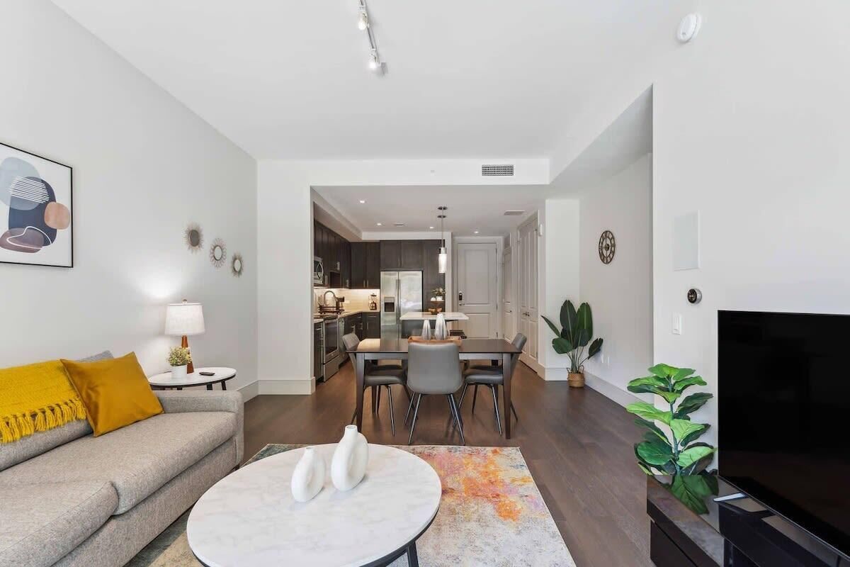 A modern living room with a sofa, a coffee table, and a TV. There is a dining area with a table and chairs. The room has a kitchen in the background.