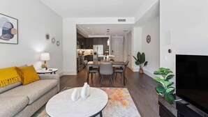 Una sala moderna con sofá, una mesa de centro y televisor. Se observa un comedor con mesa y sillas. En el fondo, hay una cocina.