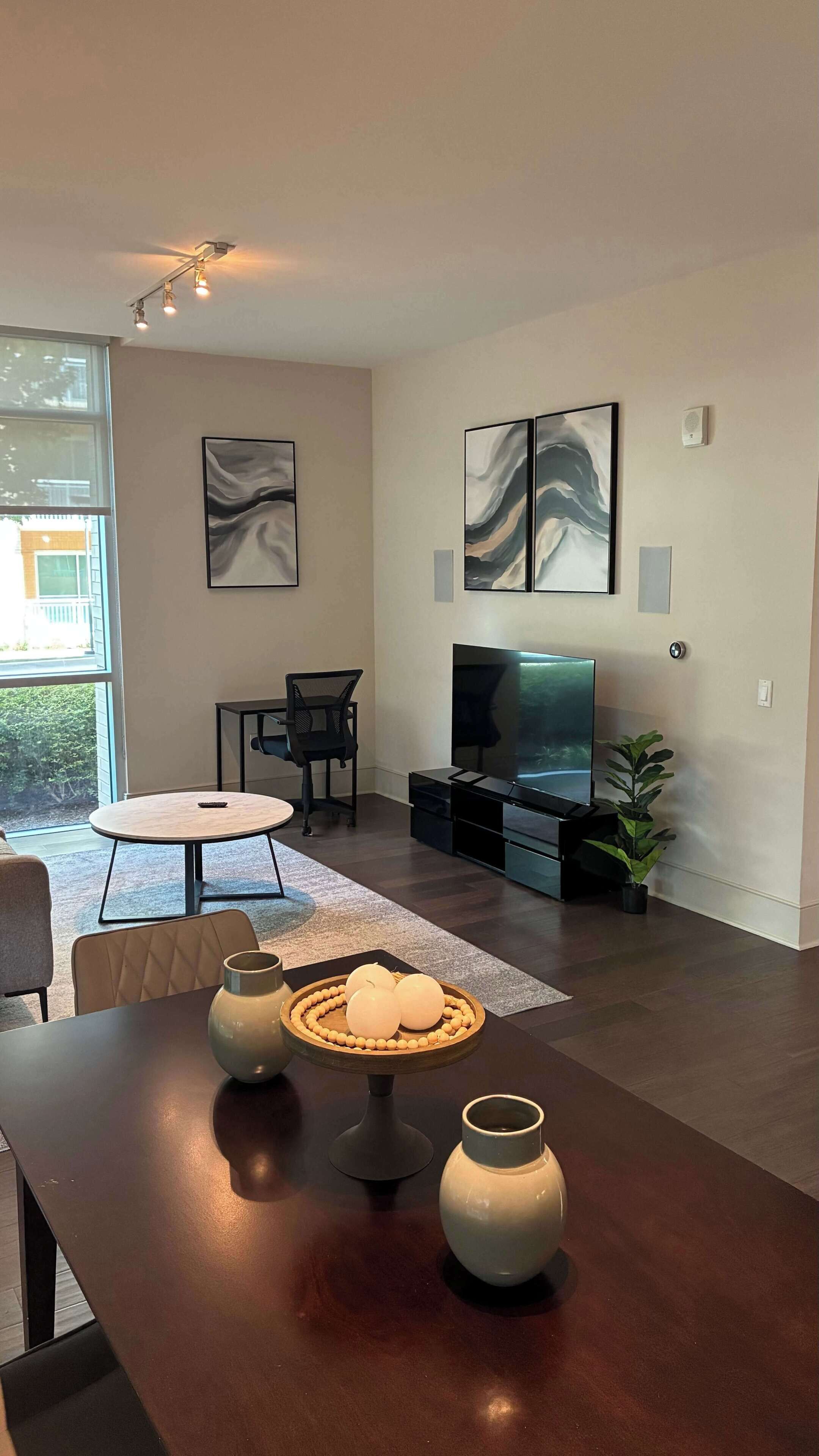 A modern living room with a flat-screen TV, a dining table with decorative items, and a large window providing natural light.