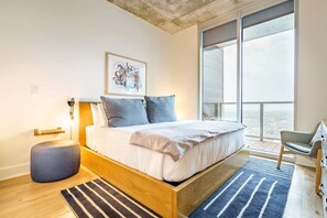Une chambre moderne avec un grand lit, une table de chevet, une chaise, une fenêtre donnant sur un beau paysage et une œuvre d’art murale.