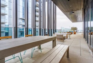Un espace extérieur moderne, aménagé avec une longue table en bois et des bancs, de grandes fenêtres et une vue sur un immeuble de grande hauteur.