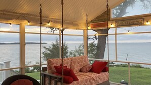 Ein überdachter Außenbereich mit einer Couch, Sesseln, einem Glastisch und Blick auf das Wasser.