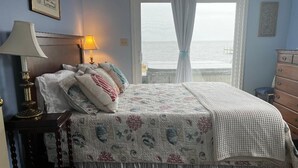 Ein Schlafzimmer mit Bett, Nachttisch, Lampe, Kommode und Meerblick durch ein Fenster.