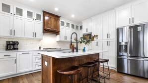 A modern kitchen with white cabinets, a stainless steel refrigerator, a wooden island with stools, and a coffee machine.