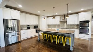 A modern kitchen with white cabinets, stainless steel appliances, a granite countertop, and a central island with yellow stools. - Spacious Lake Norman Waterfront with hot tub, game room, kayaks & SUPs! (Mooresville)