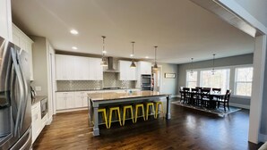A spacious kitchen with white cabinets, a central island with yellow stools, and a dining area with a dark wood table and chairs. - Spacious Lake Norman Waterfront with hot tub, game room, kayaks & SUPs! (Mooresville)