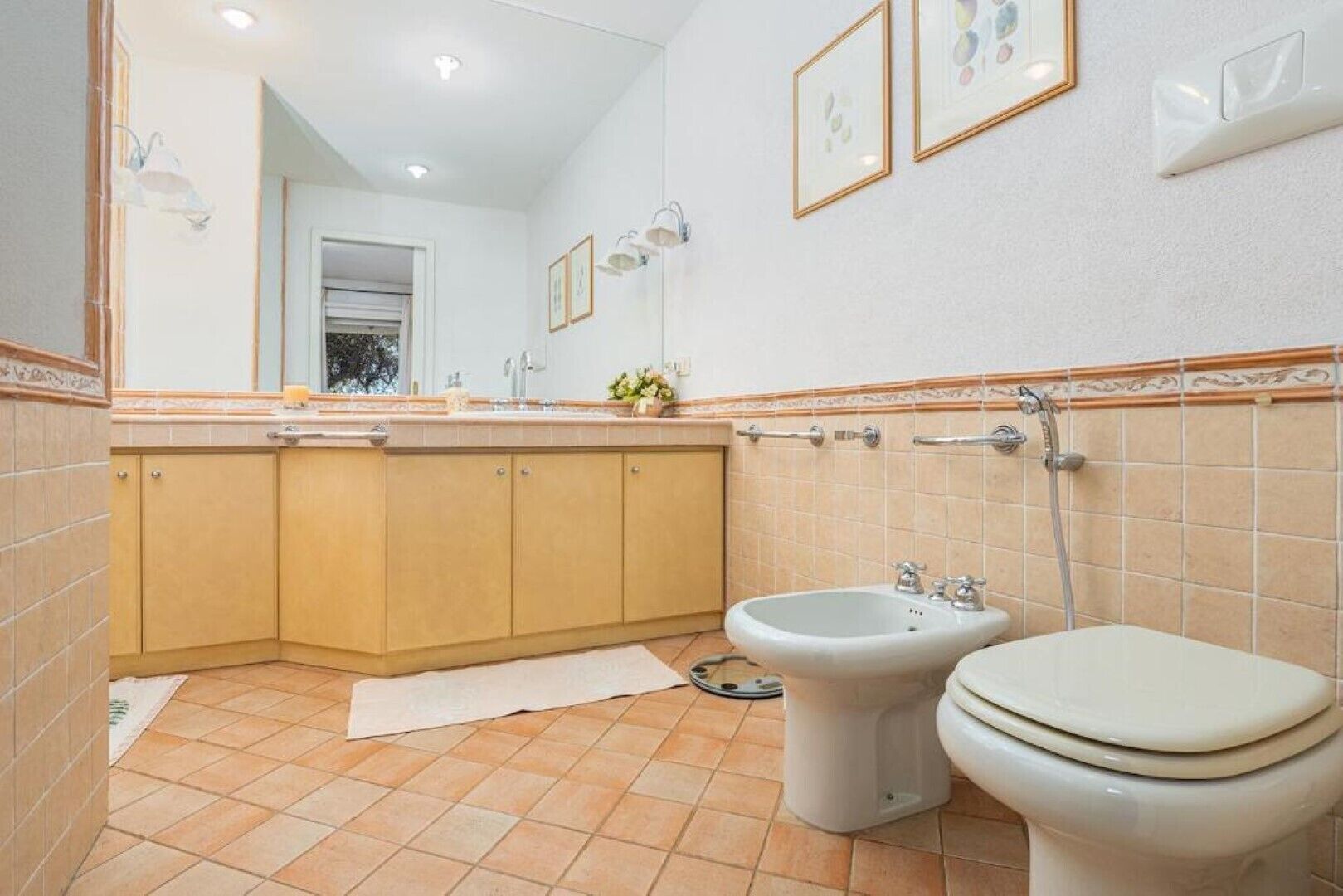 A bathroom with a sink, bidet, and toilet. There are framed pictures on the wall and a towel on the floor.