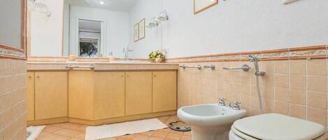 A bathroom with a sink, bidet, and toilet. There are framed pictures on the wall and a towel on the floor.