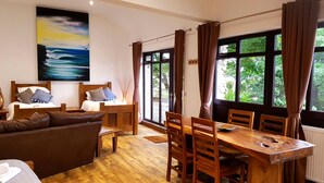 A cozy room with a bed, a wooden dining table, and chairs. There is a painting on the wall and a window with curtains.