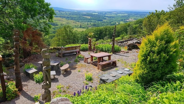 A rustic outdoor seating area with stone benches and a wooden table, surrounded by lush greenery and a scenic view of rolling hills.