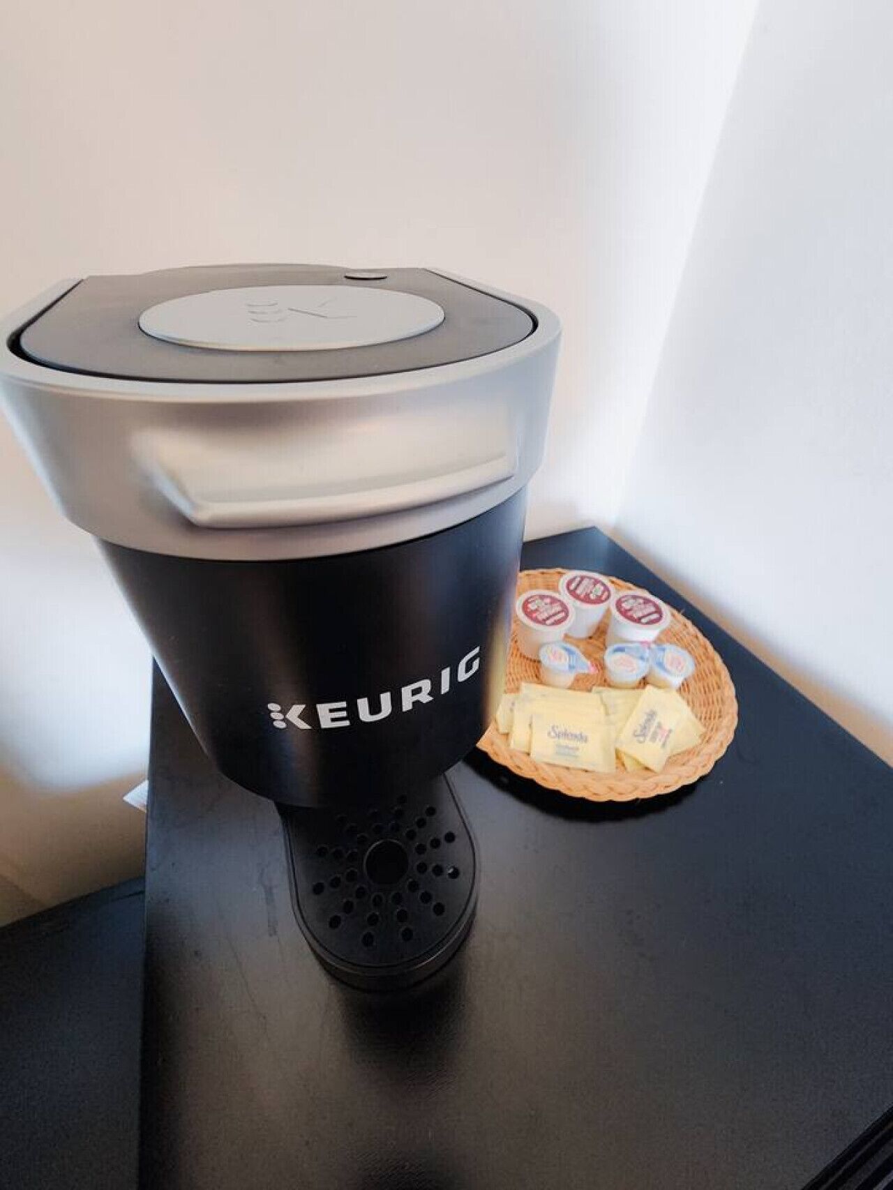 En Keurig-kaffemaskine på en sort bordplade med en kurv fyldt med kaffekapsler og en lille tallerken med ost.