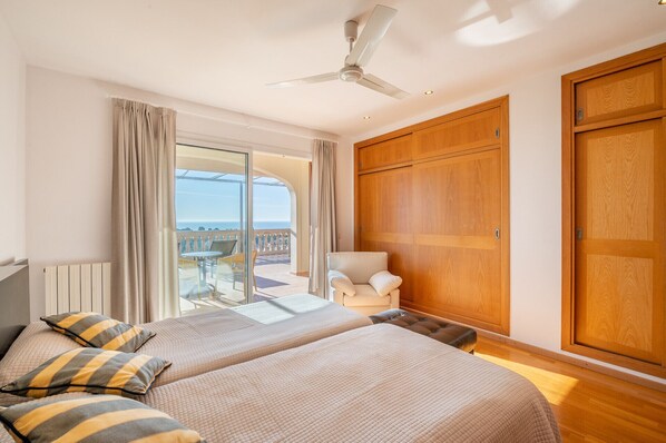 A hotel room with two beds, a ceiling fan, a large wooden wardrobe, and a view of the sea through a glass door.