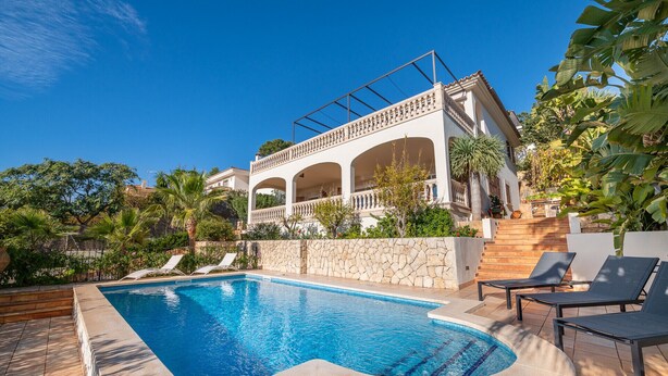 A pool area with lounge chairs, a stone wall, and a building with arches and balconies. - VILLA BENDINAT 10 - by Priority Villas (Bendinat)
