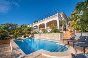 A pool area with lounge chairs, a stone wall, and a building with arches and balconies.