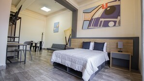 Quarto de hotel moderno com cama, mesas de cabeceira, escrivaninha com cadeiras e área de jantar.