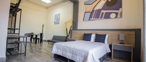 A modern hotel room with a bed, bedside tables, a desk with chairs, and a dining area.