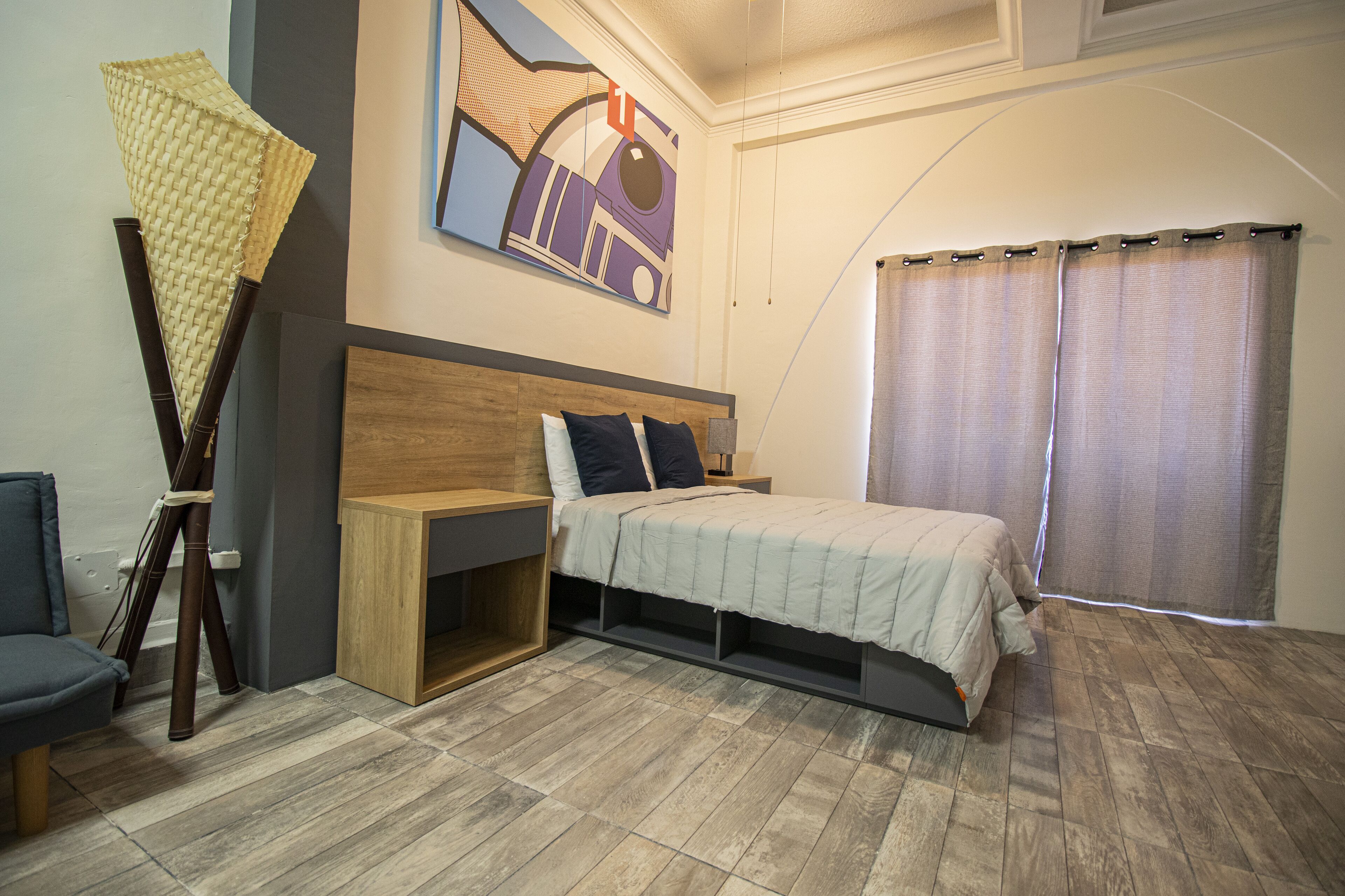 A bedroom with a bed, a wooden nightstand, a desk, a chair, and a large window with curtains.