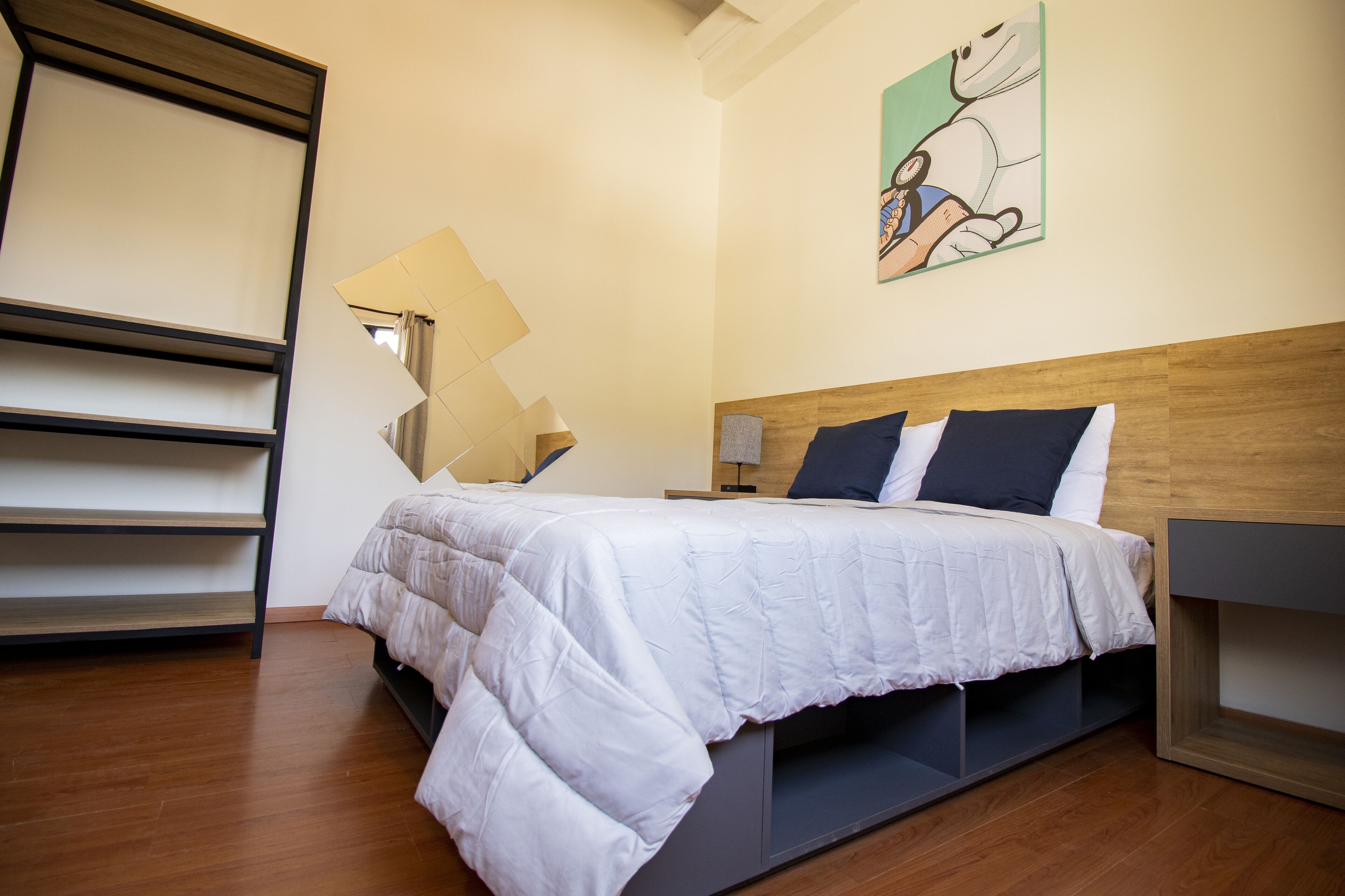 Un dormitorio con una cama, una mesita de noche, una lámpara de pared, un cuadro y suelo de madera.
