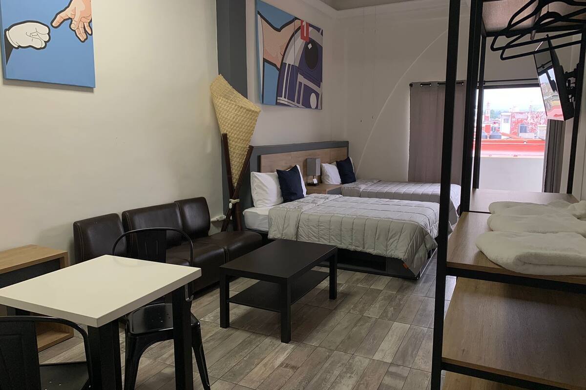A hotel room with two beds, a sofa, a table, and a chair. There is a painting on the wall and a window with a view of buildings.