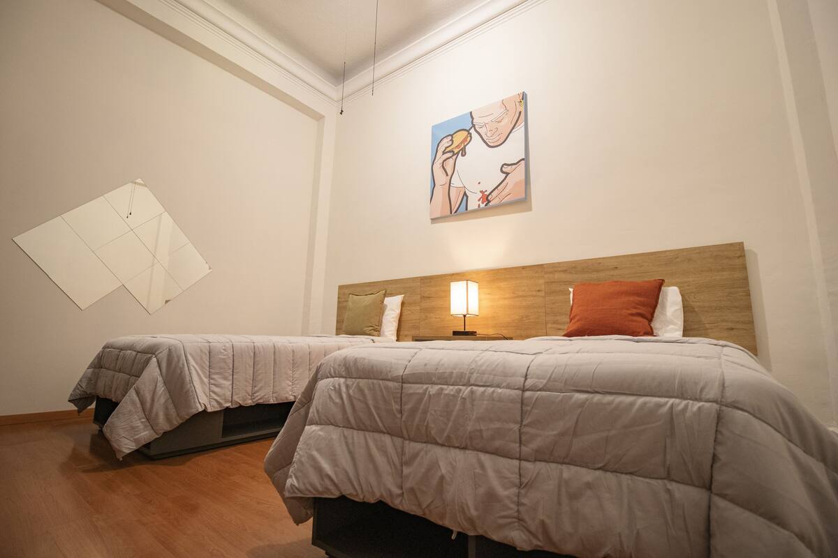 A hotel room with two beds, a wooden headboard, a wall-mounted artwork, and a floor lamp.