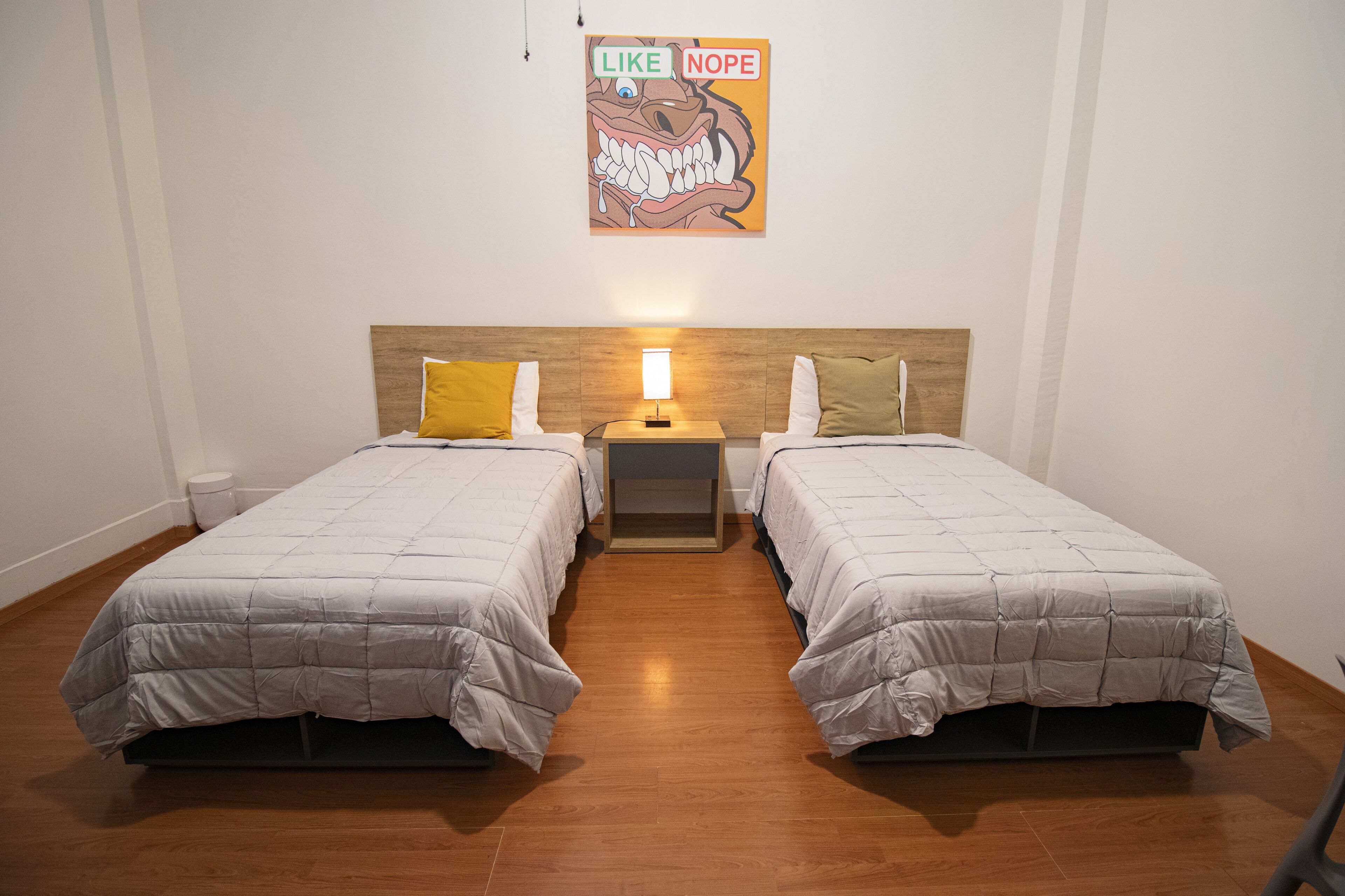 Habitación de hotel con dos camas individuales, cabecera de madera, mesita de noche con lámpara y un cartel en la pared.
