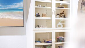 A white shelving unit with books and decorative items, a framed painting of a beach, and a white lamp.