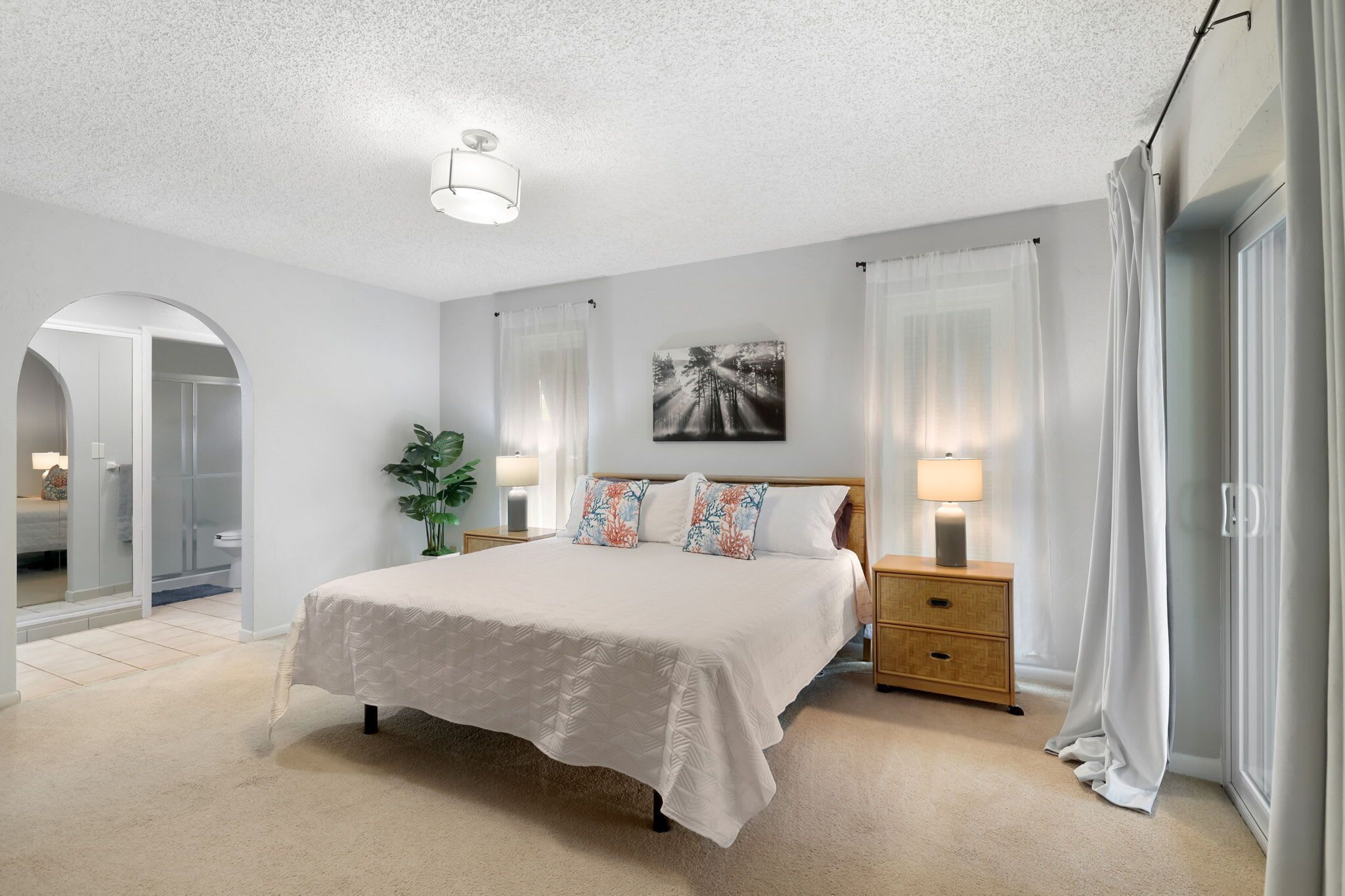 Quarto com cama, mesas de cabeceira, luminária, planta e uma porta com cortinas.