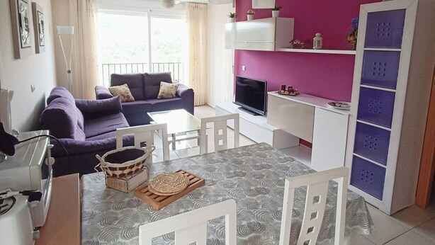 Een moderne woonkamer met een paarse bank, een eettafel met stoelen, een televisie en een keuken met een roze muur en kastjes.