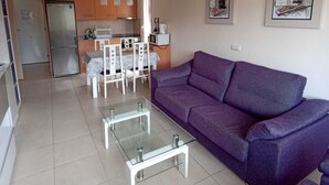 Een moderne woonkamer met een paarse bank, een glazen salontafel en een eethoek met een witte tafel en stoelen.