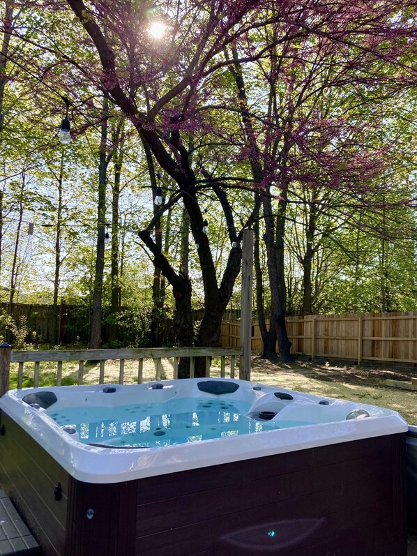 Un jacuzzi en un patio trasero con una terraza de madera y árboles al fondo.
