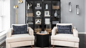 Ein Wohnzimmer mit zwei Sesseln, einem Couchtisch, einem gemusterten Teppich, einer schwarzen Regaleinheit mit verschiedenen Dekorationsobjekten und einer Stehlampe.