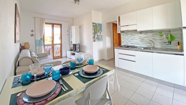 A modern kitchen with white cabinets, a gas stove, and a sink. There is a dining area with a table set for two, featuring blue glassware and plates. - Sunset House PuntoCasaBudoni (San Teodoro)