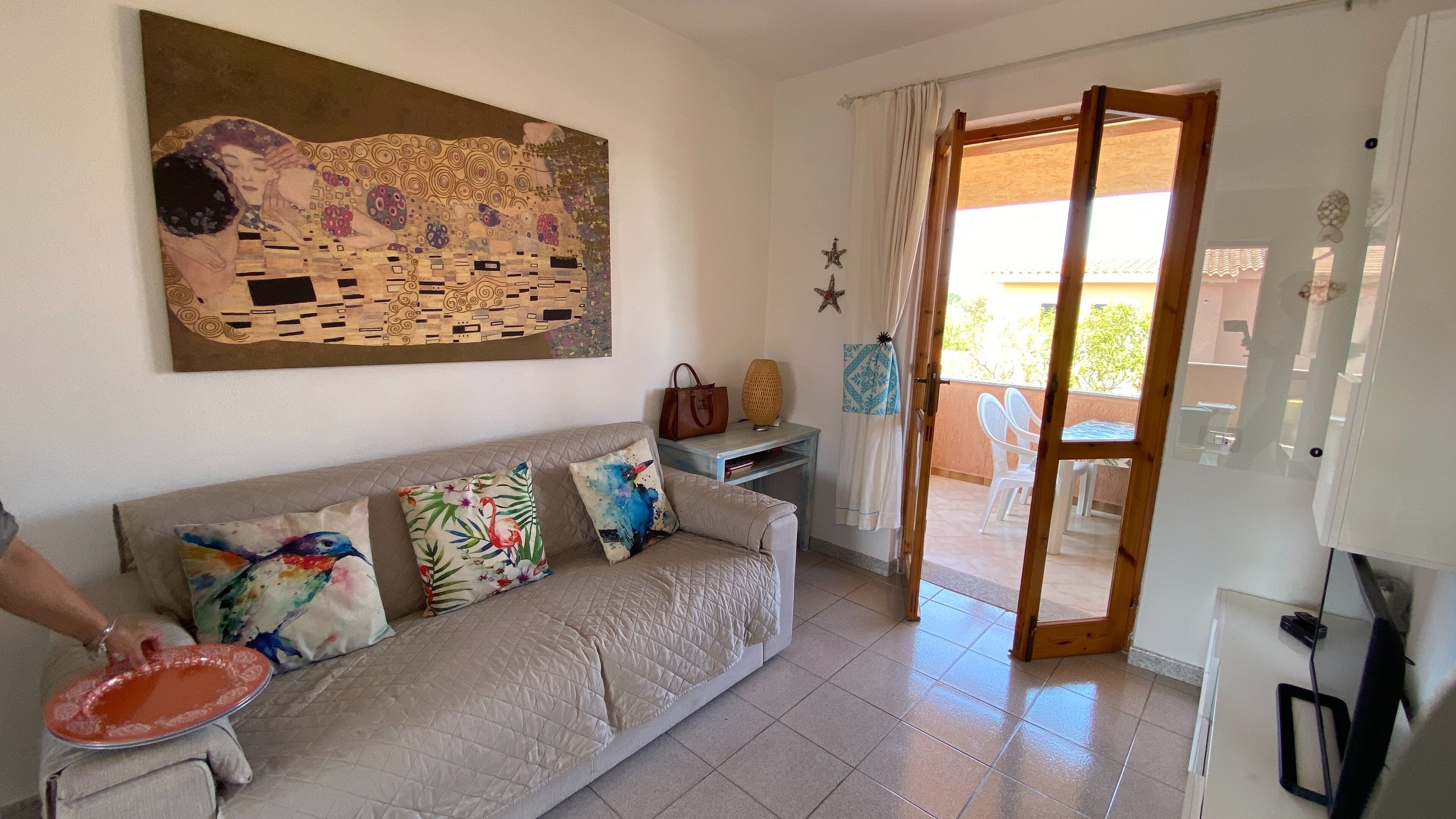 A living room with a beige sofa, colorful cushions, a painting on the wall, and a view of the outside balcony.