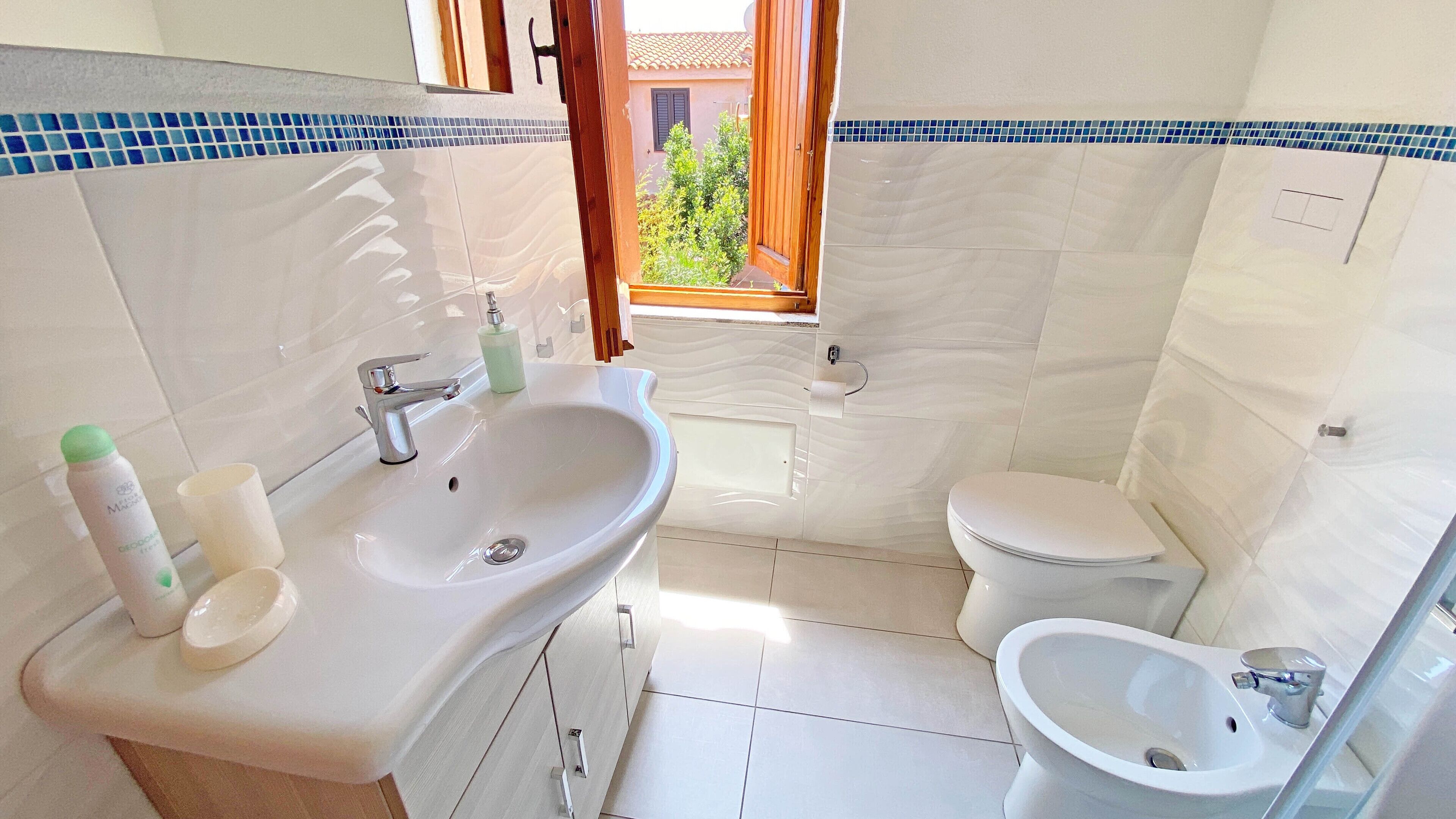 Une salle de bain équipée d’un lavabo, de toilettes, d’un bidet et d’une fenêtre donnant sur un espace verdoyant.