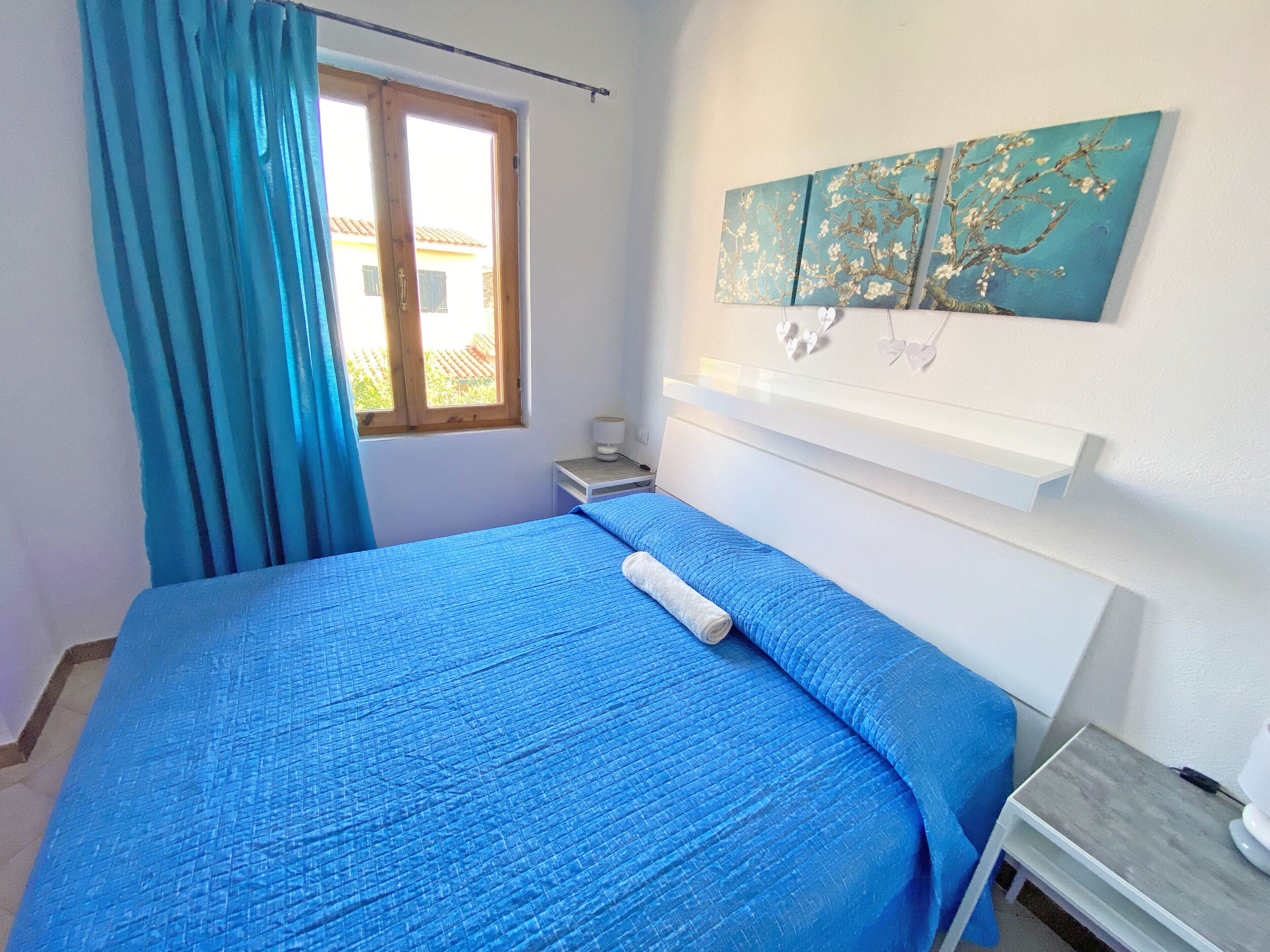 Un dormitorio con cama, mesita de noche, ventana con cortinas y un cuadro colgado en la pared.