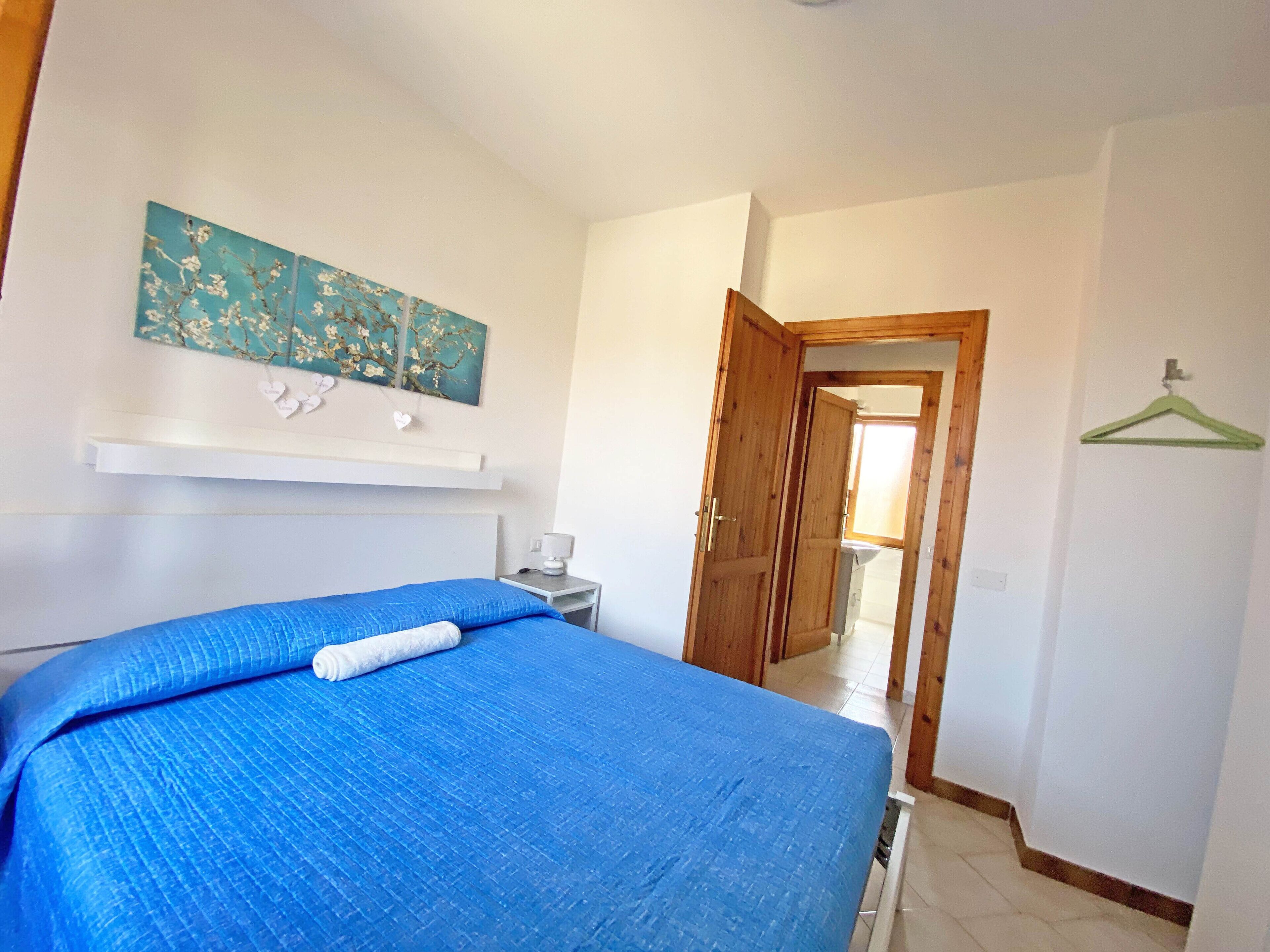 Un dormitorio con cama, mesita de noche, un cuadro y una puerta que da a otra habitación.