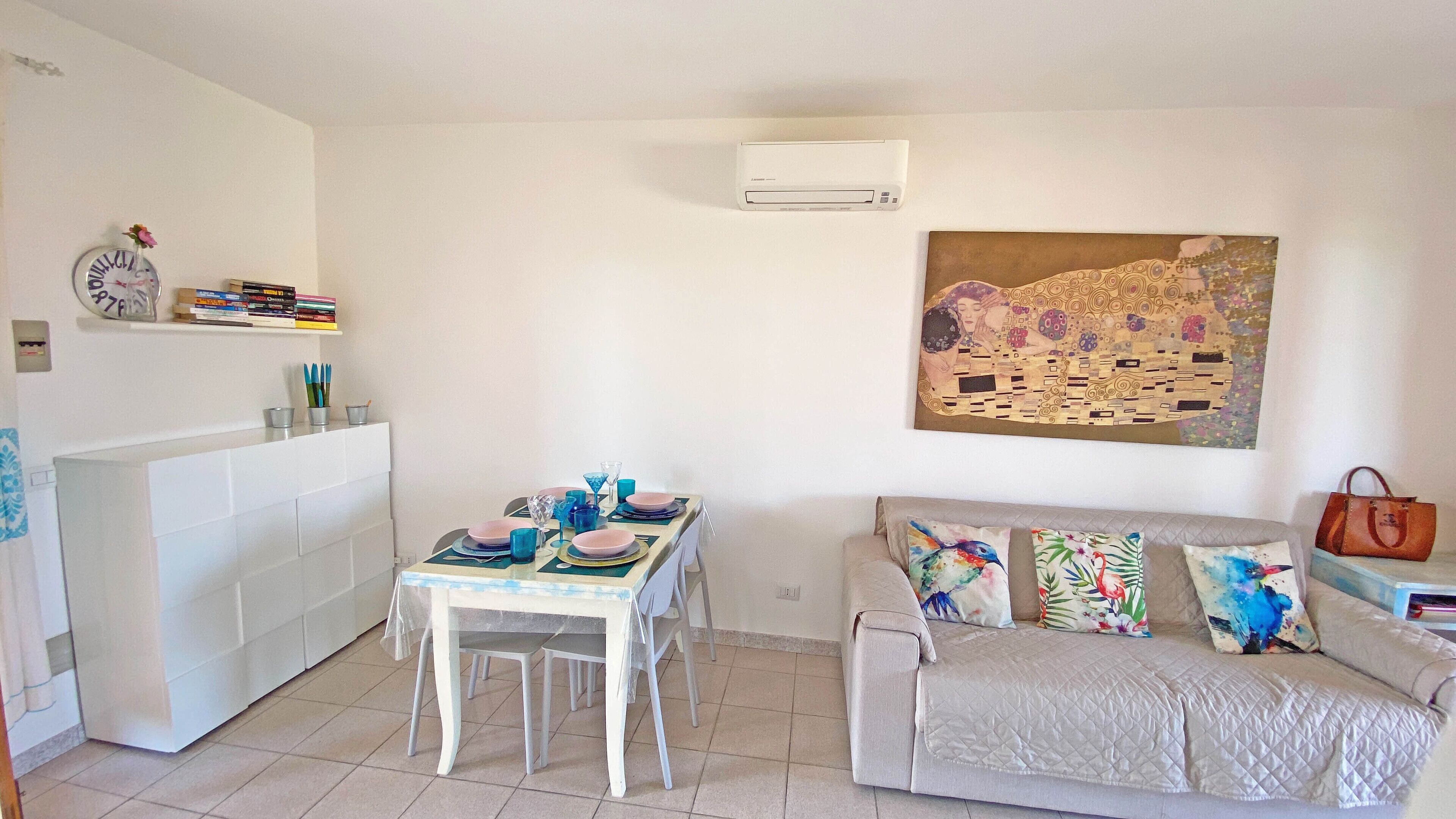 A dining area with a table set for four, a sofa with colorful cushions, a clock, and a wall-mounted air conditioner.