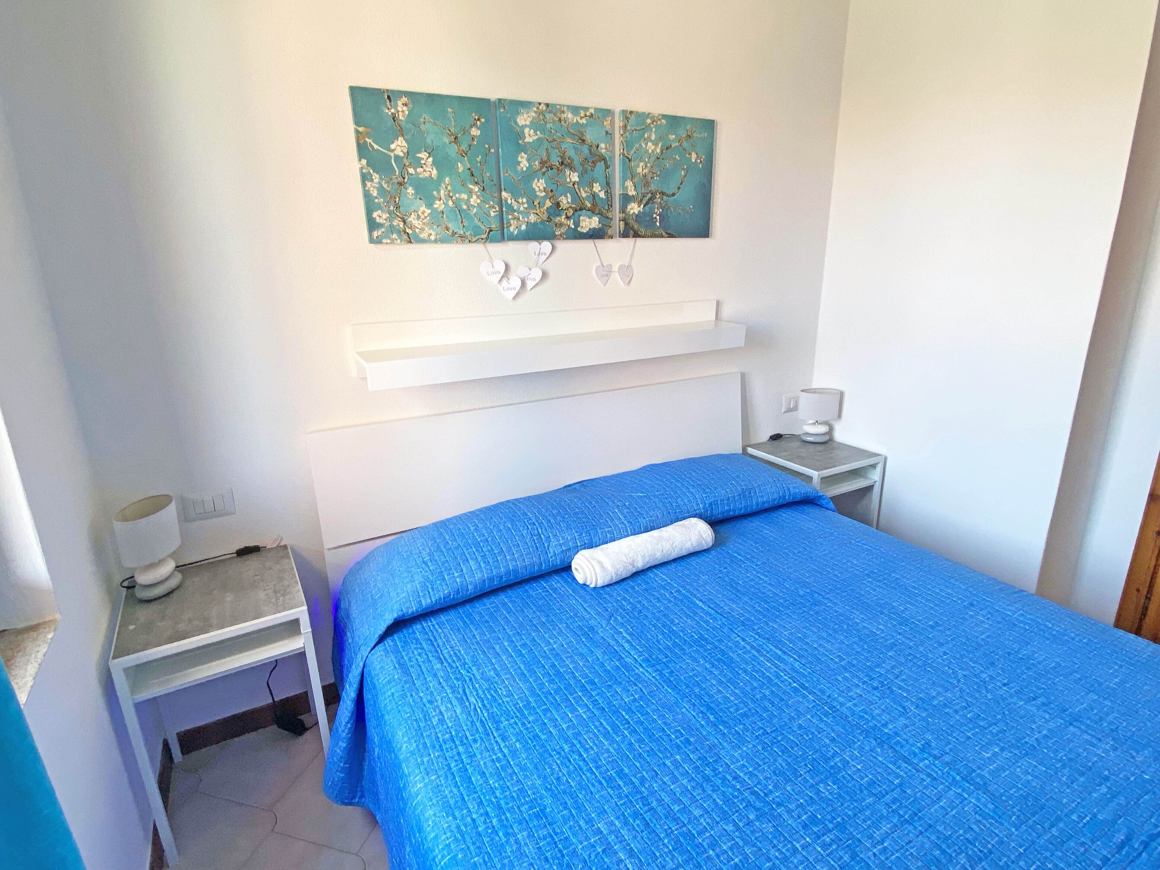 Un dormitorio con colcha azul, una mesita de noche blanca con lámpara y una estantería sobre la cama.