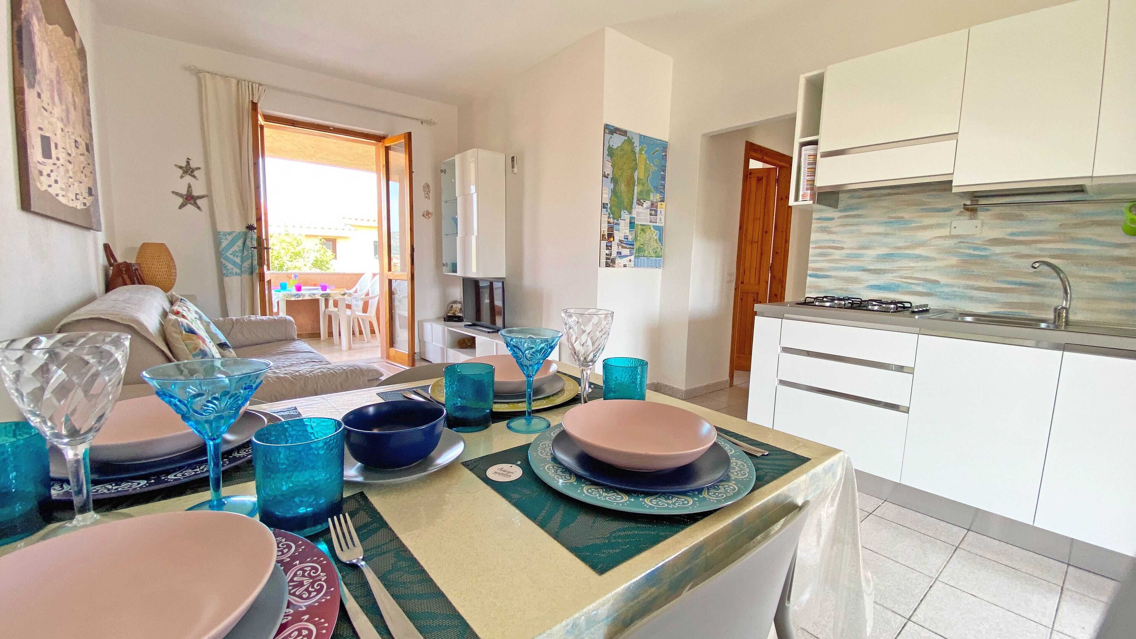 Una cocina moderna con armarios blancos, una estufa a gas y un lavamanos. Hay una zona de comedor con una mesa preparada para dos personas, que incluye vasos y platos azules.