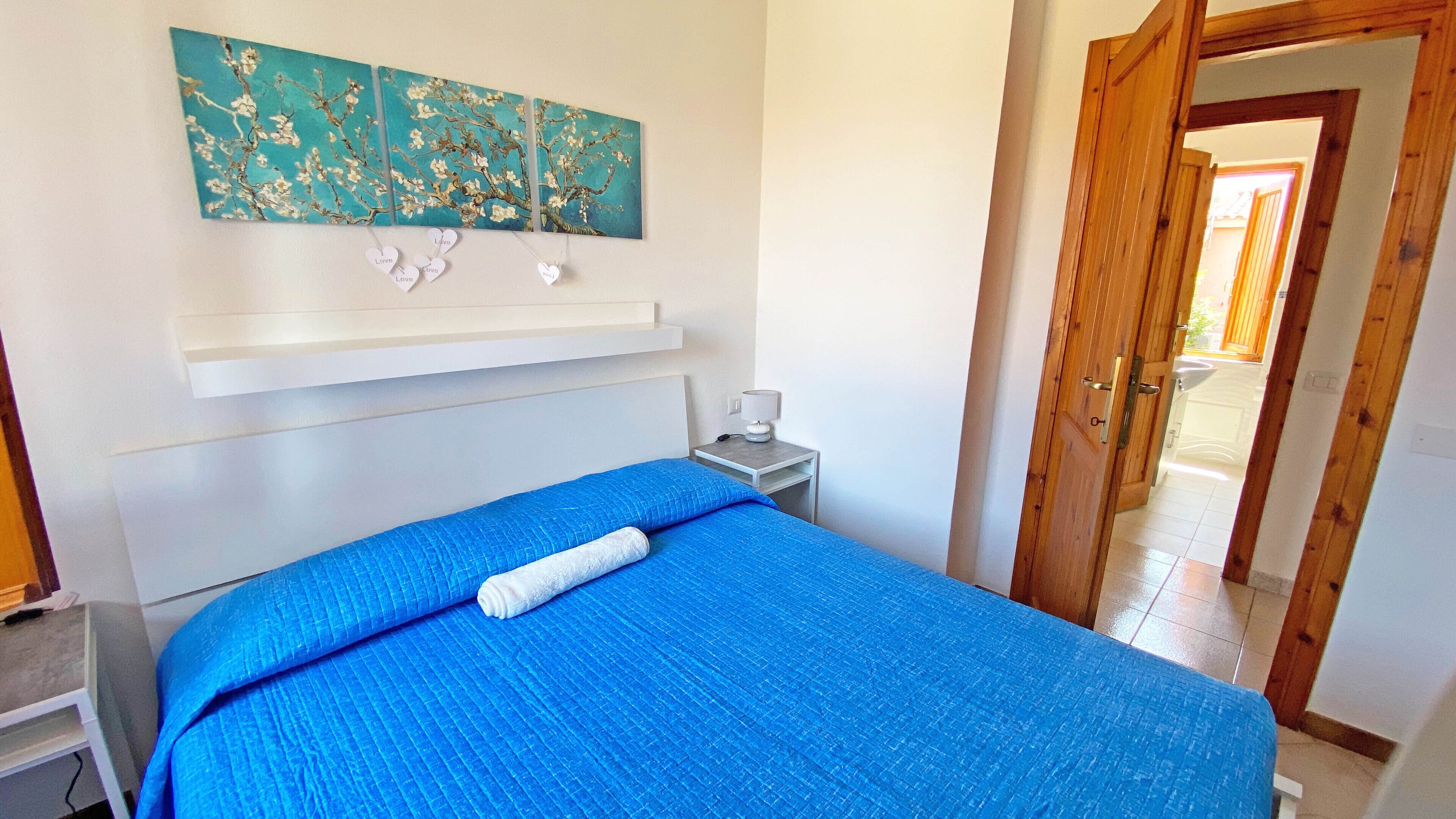 A bedroom with a bed, a nightstand, a wall-mounted shelf, and a door leading to another room.