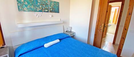 A bedroom with a bed, a nightstand, a wall-mounted shelf, and a door leading to another room.