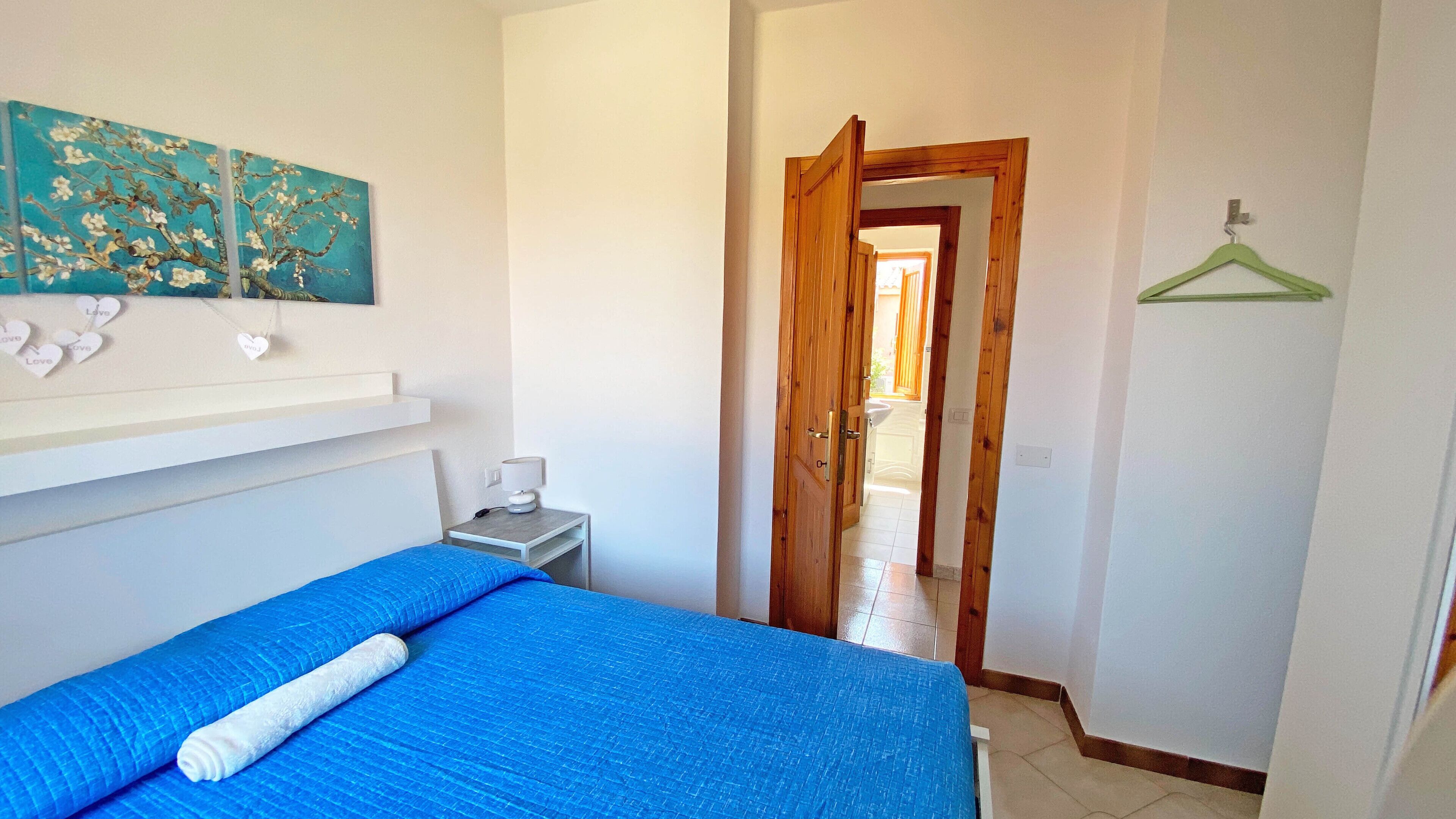 Um quarto com cama, mesa de cabeceira, prateleira na parede com objetos decorativos e uma porta que dá para outro cômodo.