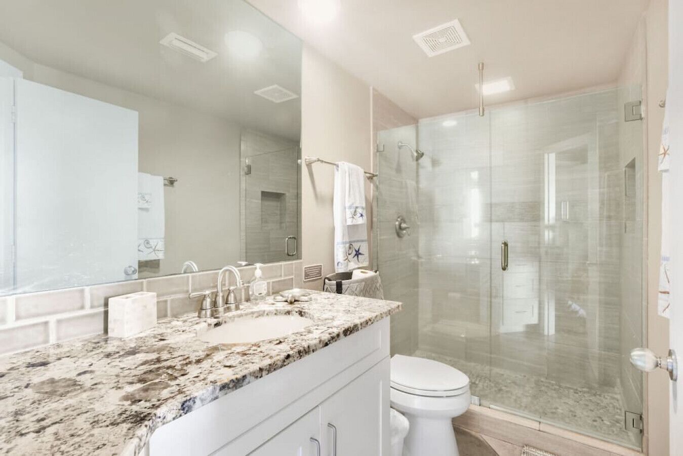 A modern bathroom with a granite countertop, a large mirror, a glass shower enclosure, and a white toilet.
