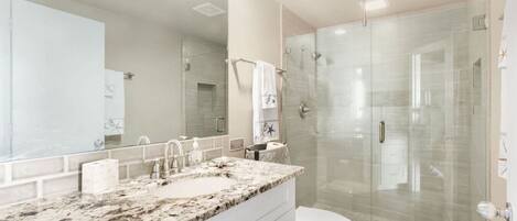 A modern bathroom with a granite countertop, a large mirror, a glass shower enclosure, and a white toilet.